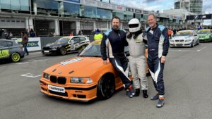 Das Fahrerteam steht mit dem orangefarbenen BMW M3 E36 in der Startaufstellung vor der Boxengasse am Nürburgring.